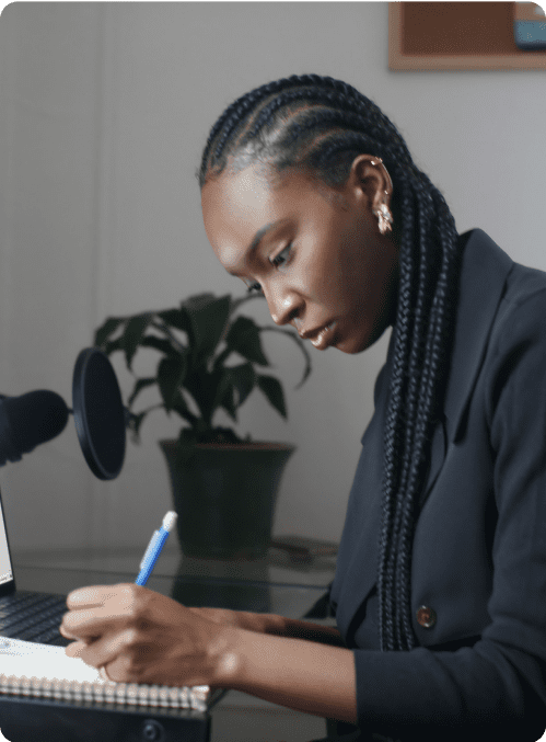 A woman, alone, writing in a notepad with a plant in the background.